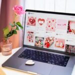 A stylish workspace featuring a laptop displaying vibrant Valentine's Day images, next to a single pink rose in a vase.
